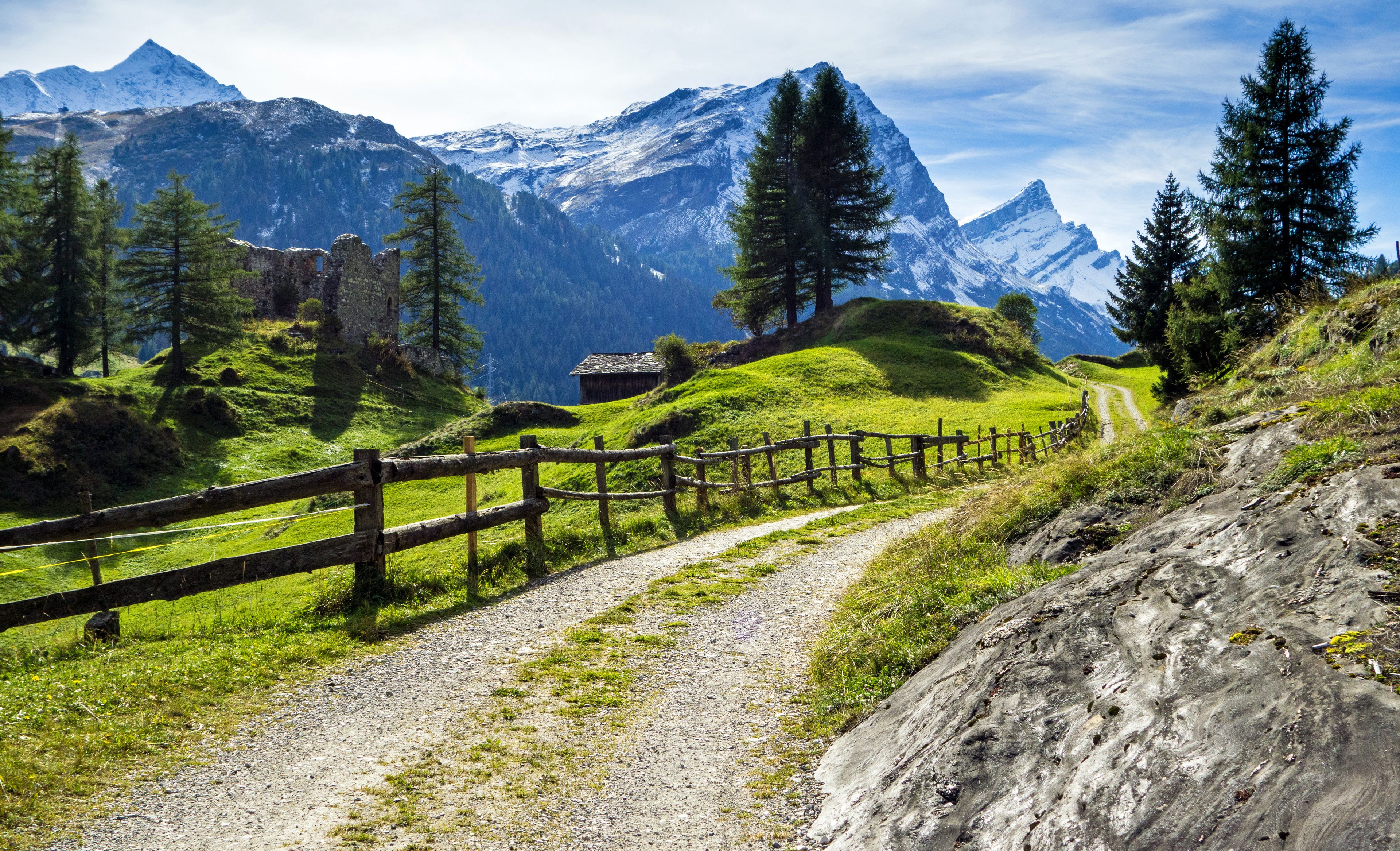 Feldweg zwischen grünen Wiesen und Ruinen, mit den Alpen im Hintergrund