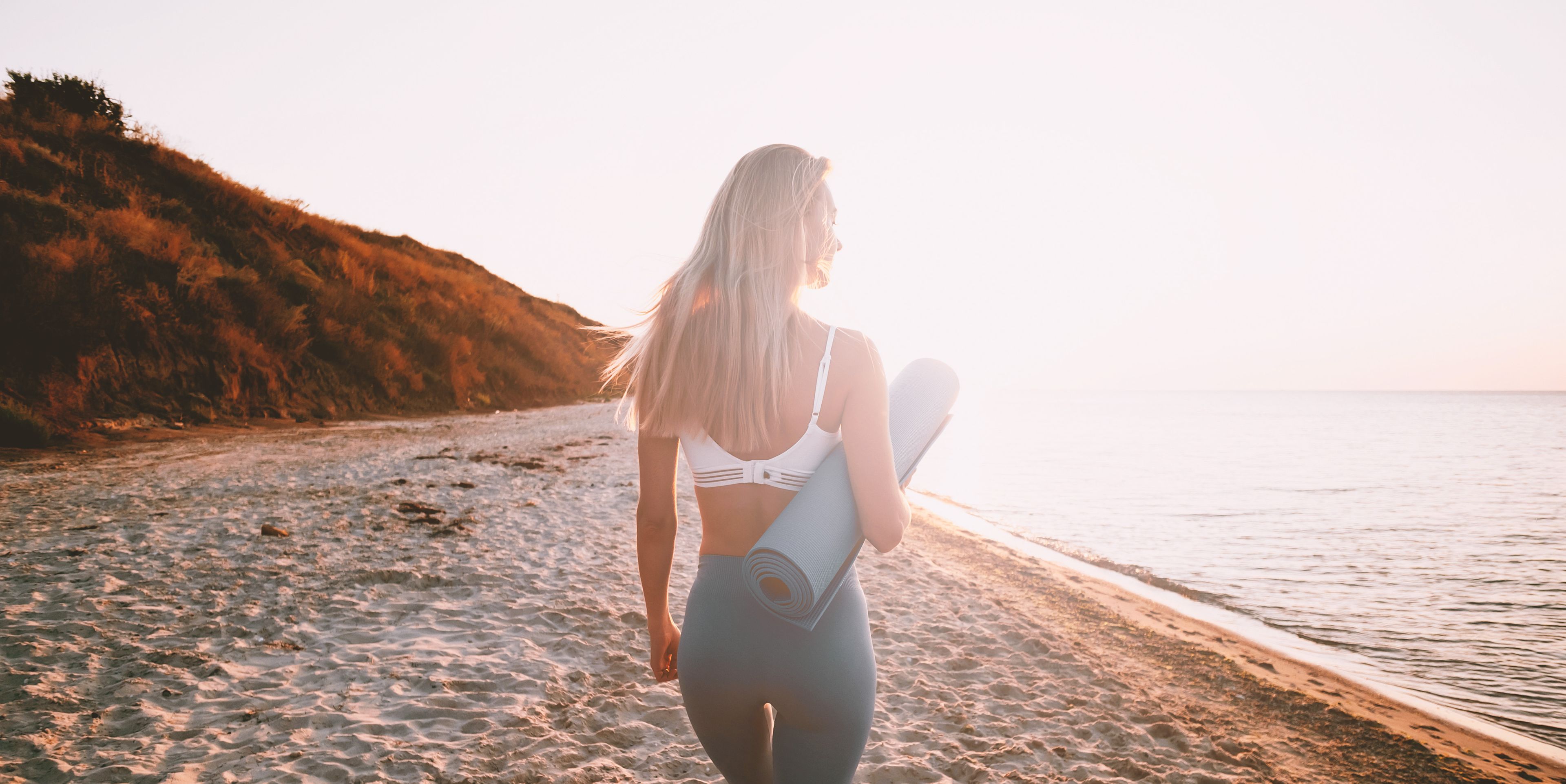 Frau mit Yogamatte unter dem Arm geht am Strand der Sonne entgegen