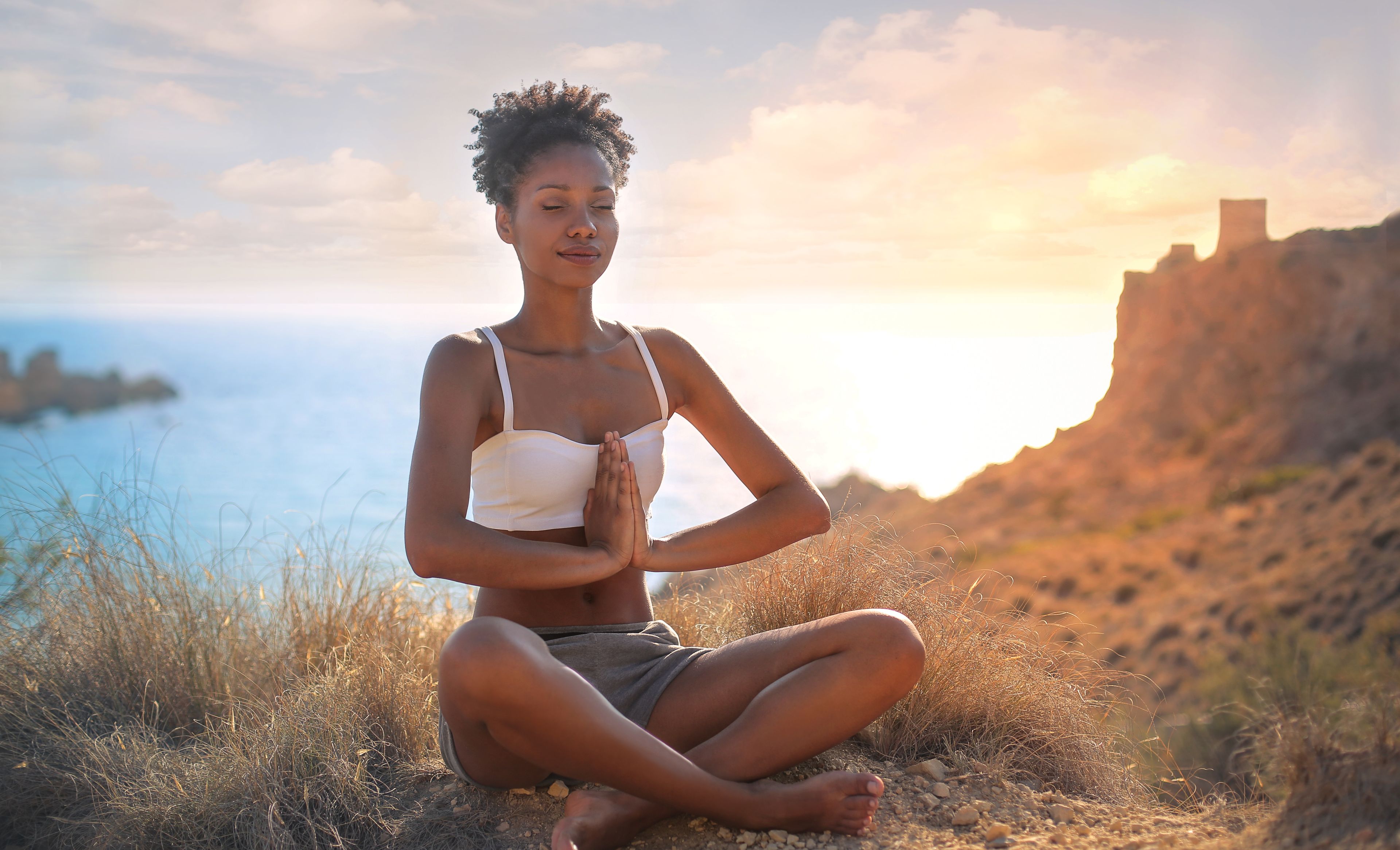 Eine Frau im Schneidersitz auf einem Felsen, die Hände vor der Brust gefaltet, im Hintergrund das Meer und der Sonnenuntergang.