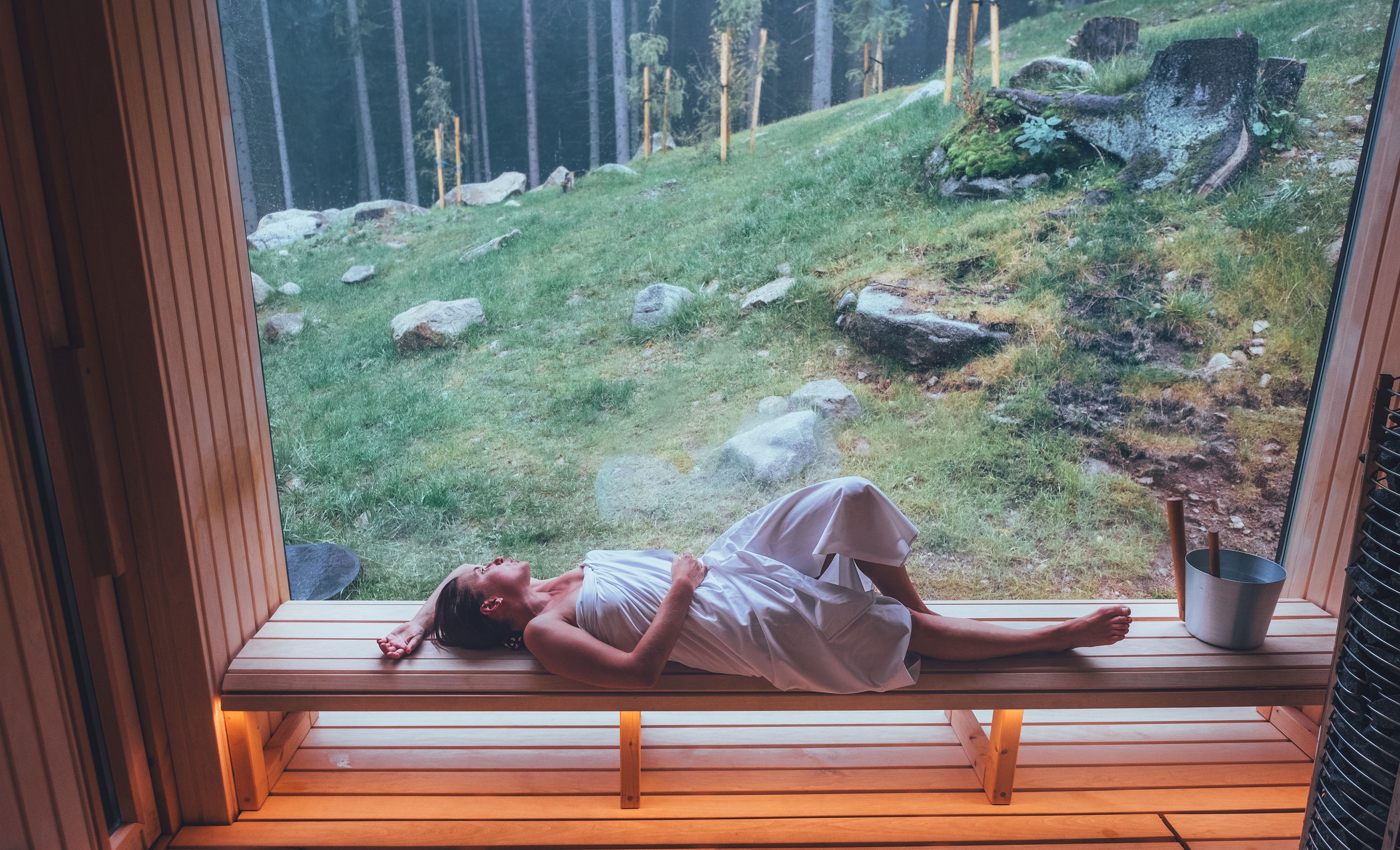 Eine Frau liegt auf einer Bank in der Sauna und schaut aus einem großen Fenster in die grüne Landschaft.