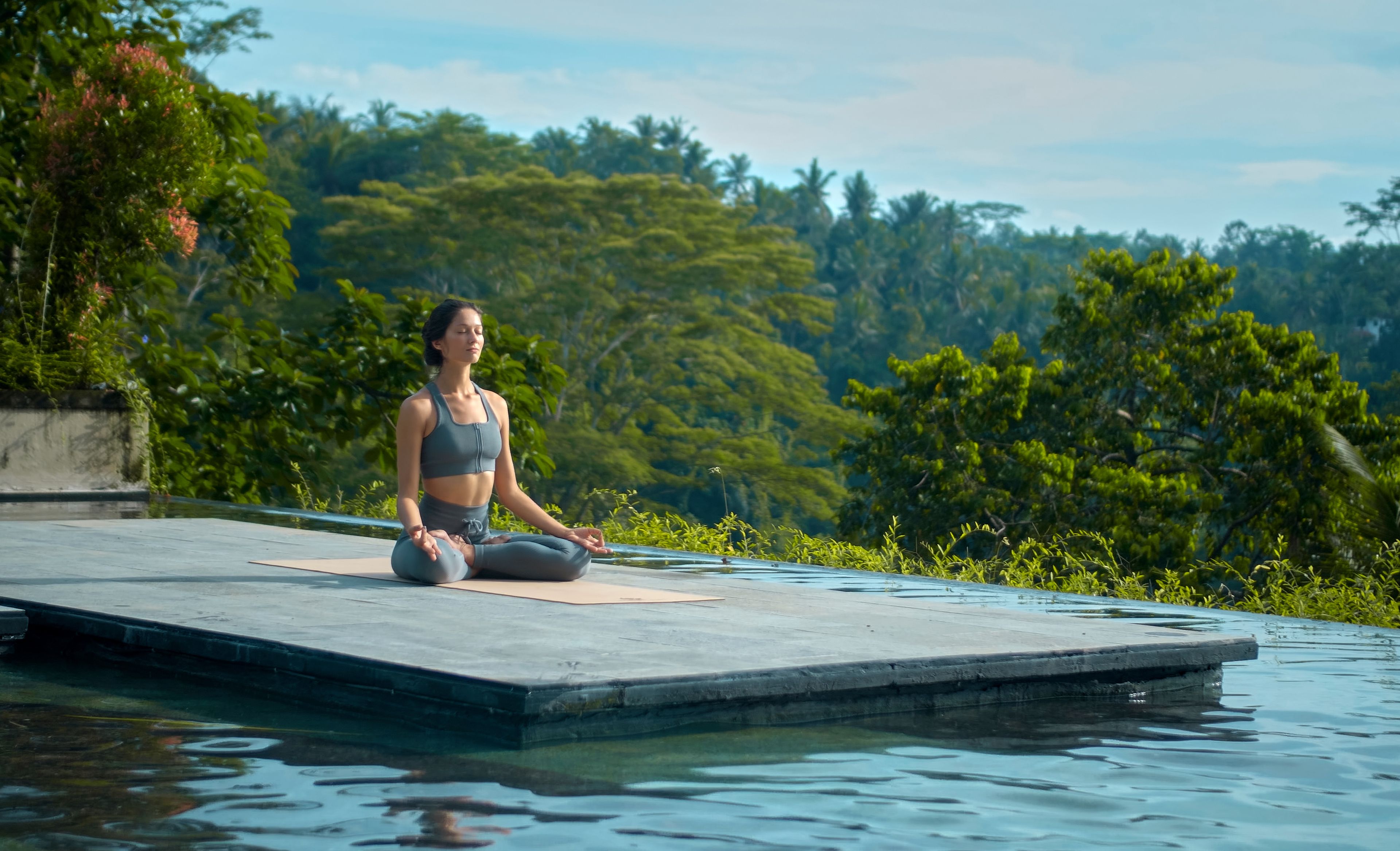 Meditierende Frau im Schneidersitz an einem Pool in tropischer Umgebung 