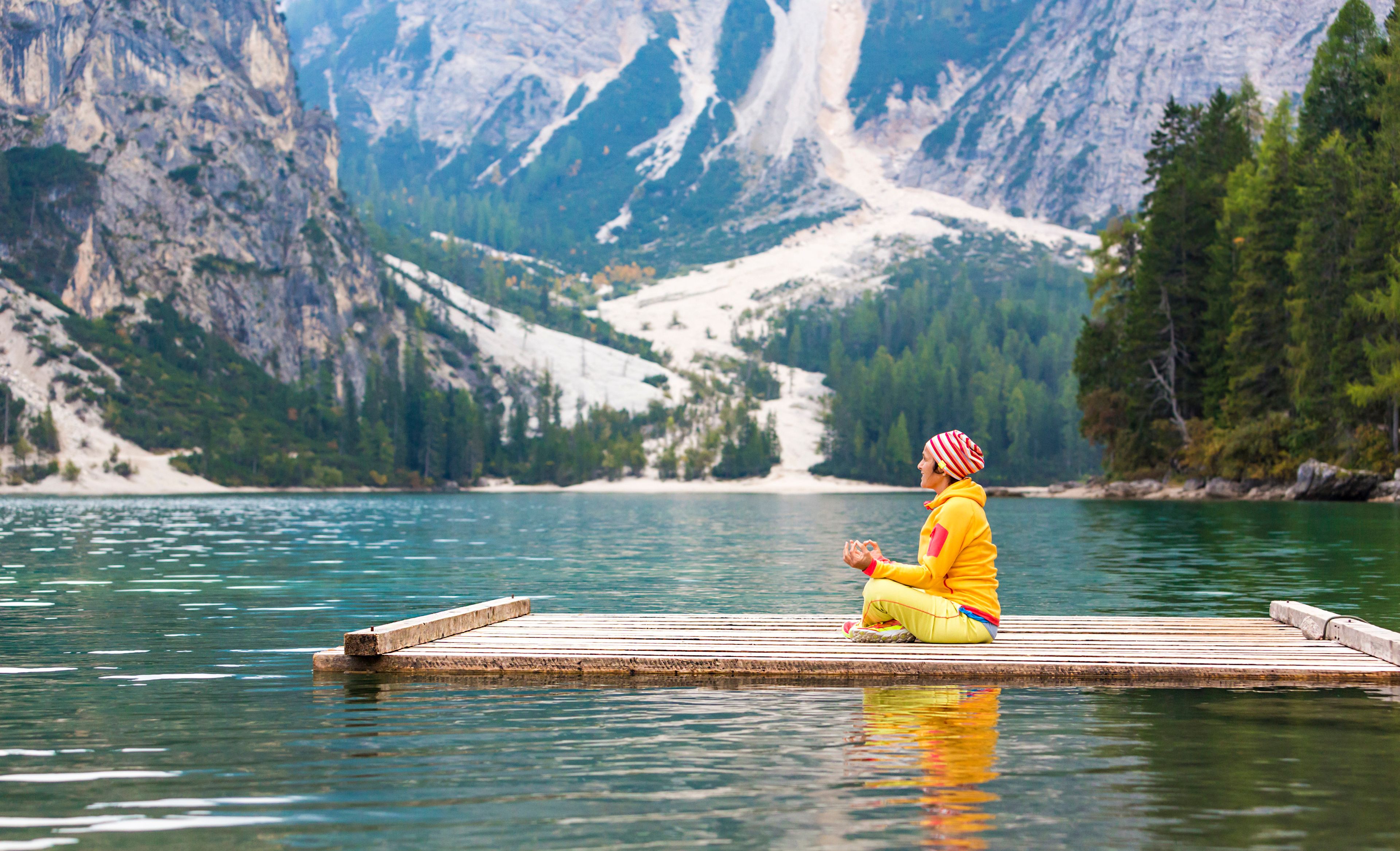 Eine Frau im Lotussitz auf dem Steg eines Sees, im Hintergrund eine beeindruckende Berglandschaft.