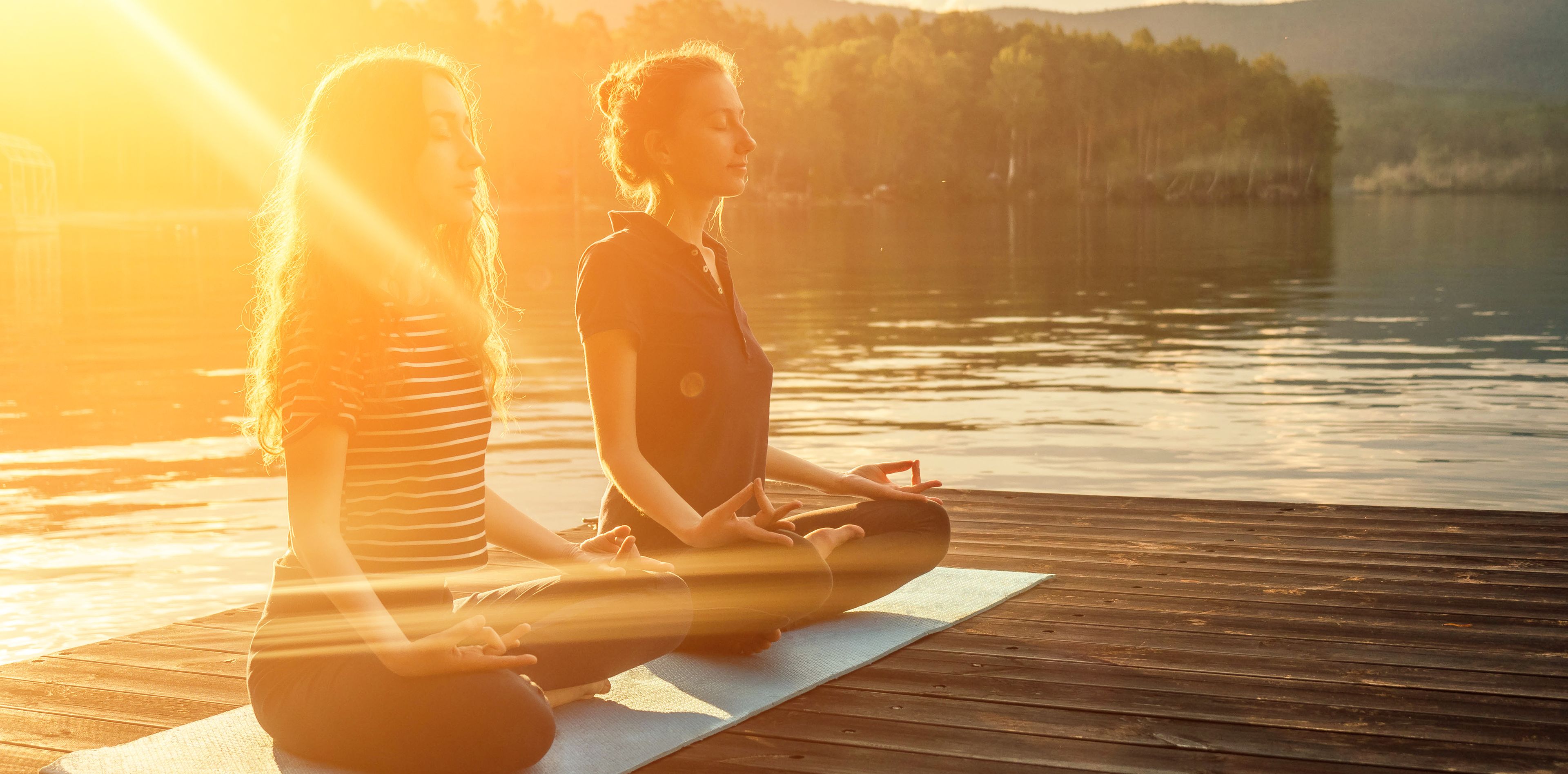 Zwei Frau meditieren auf Holzsteg am Wasser, im Sonnenschein