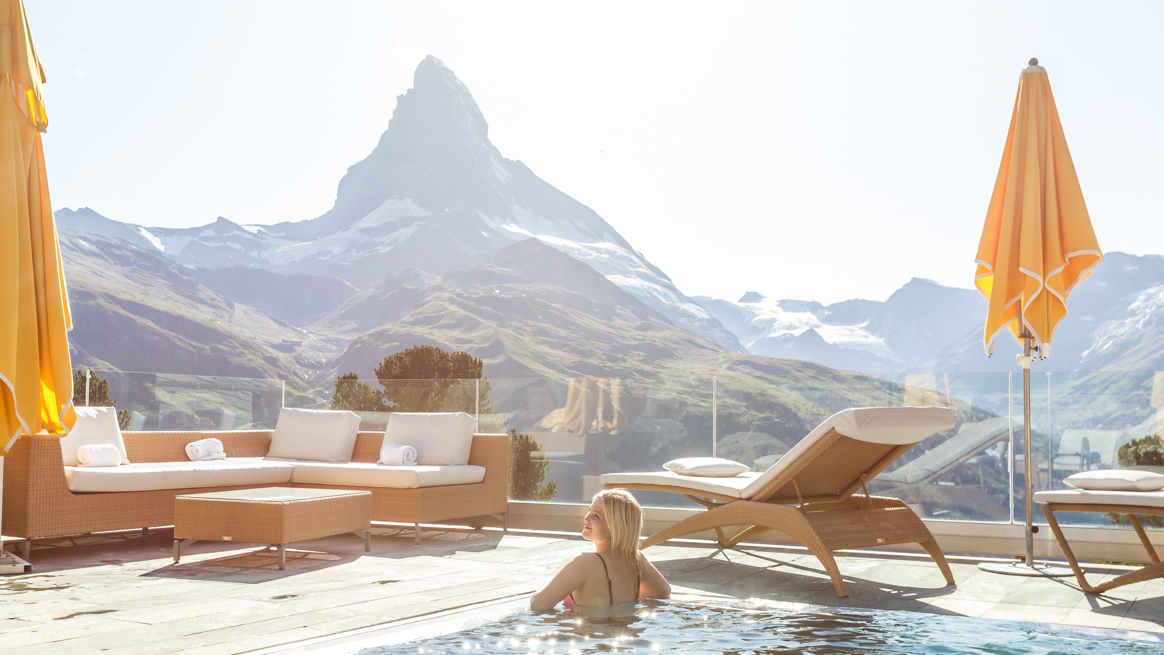Eine Frau im Pool einer Hotelterrasse die auf eine beeindruckende Berglandschaft blickt 